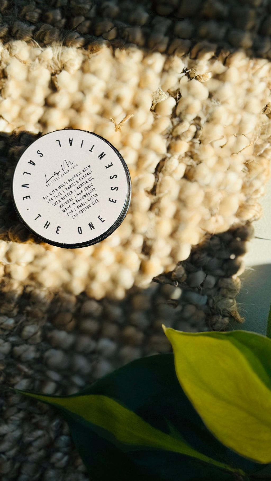 multi purpose balm with lid on, placed on a hessian rug, plant in the bottom right hand corner