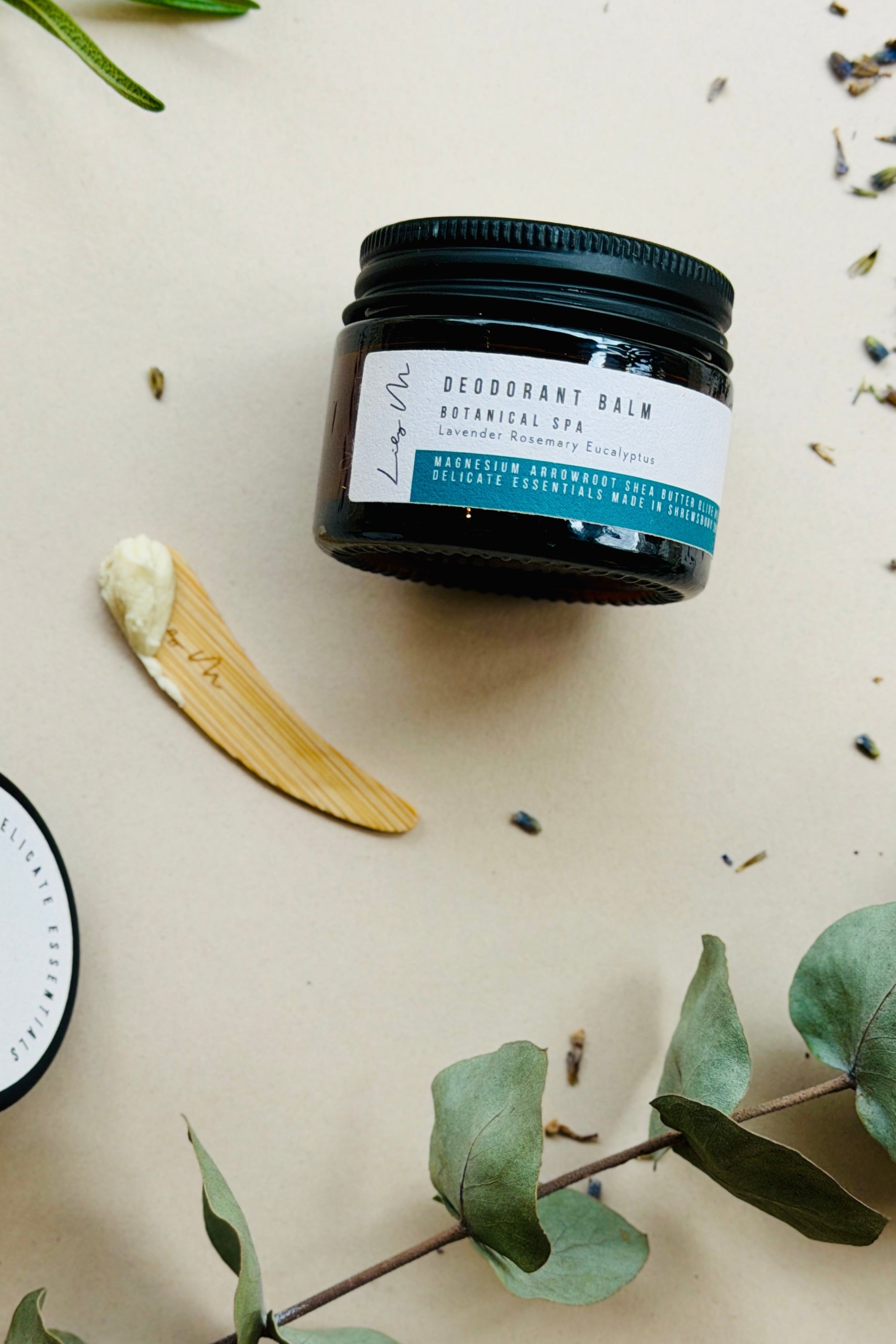 Deodorant balm jar with a wooden applicator on a light background with green leaves.