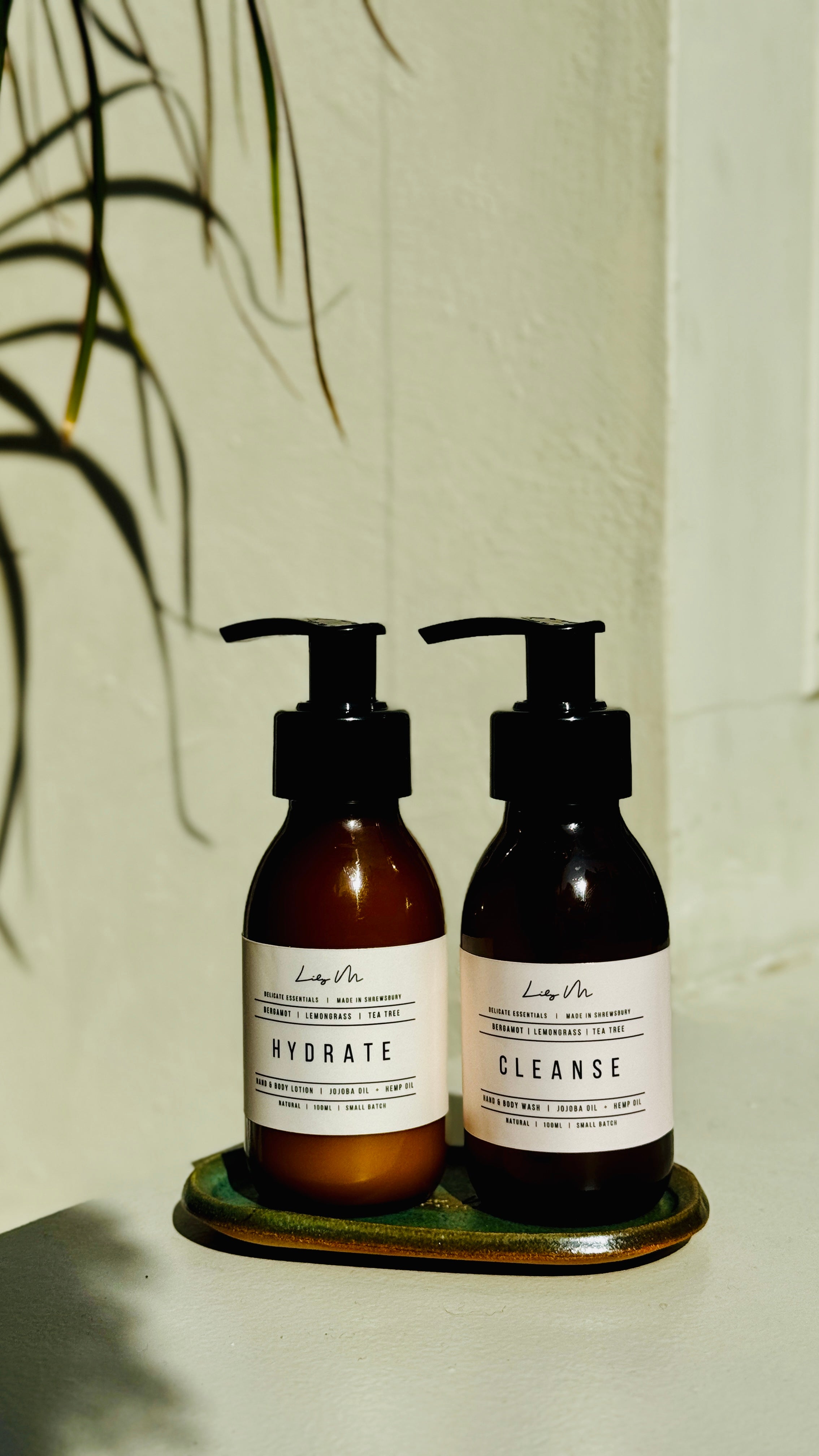 Two brown bottles with pump dispensers labeled 'Hydrate' and 'Cleanse' on a white surface with a plant in the background.