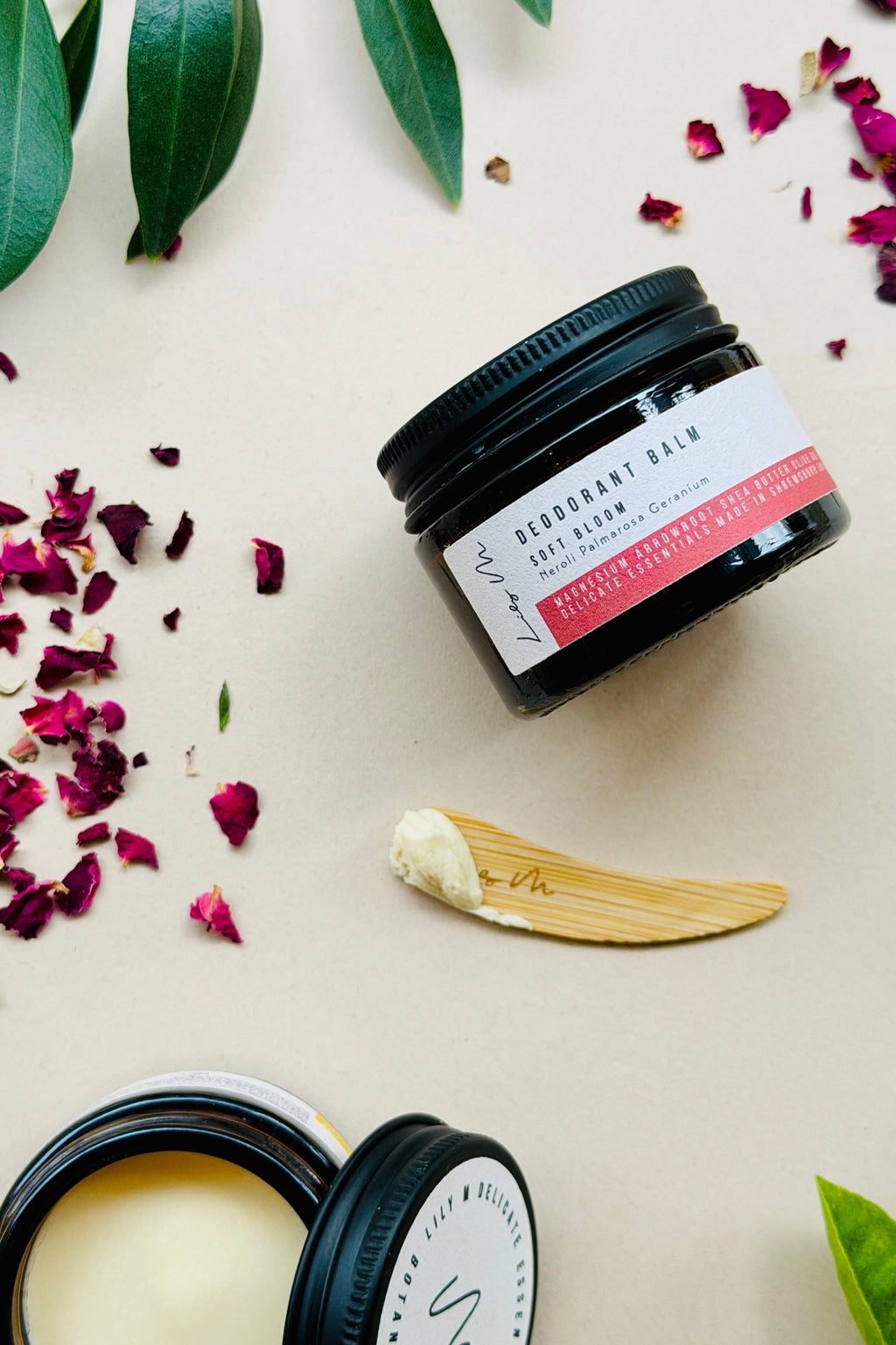 Two black jars of deodorant cream with a wooden applicator on a light background with green leaves and pink petals.